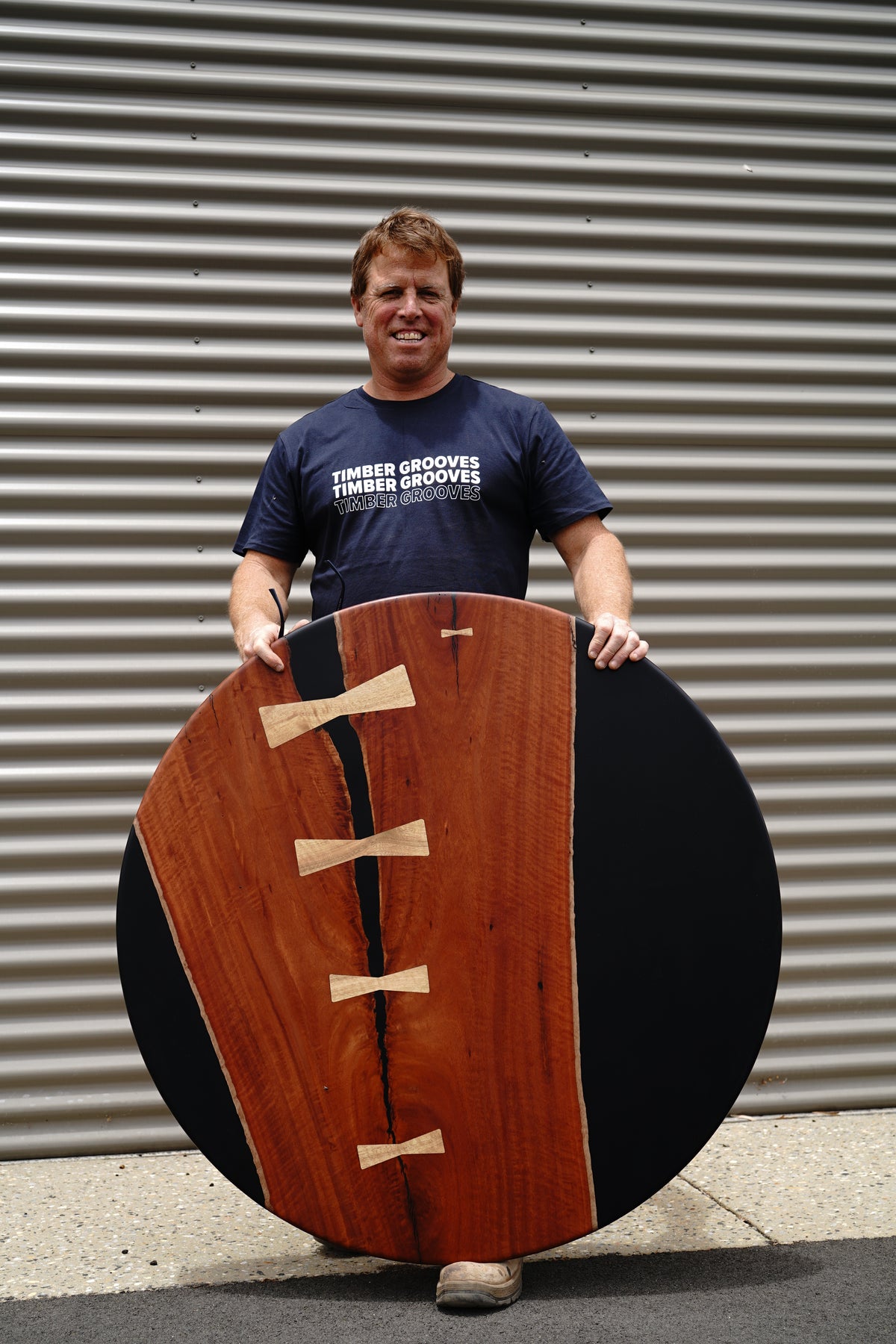 Person standing outdoors holding a Jarrah Tuxedo Coffee Table by Timber Grooves, wearing a "Timber Grooves" T-shirt, against a corrugated metal wall.