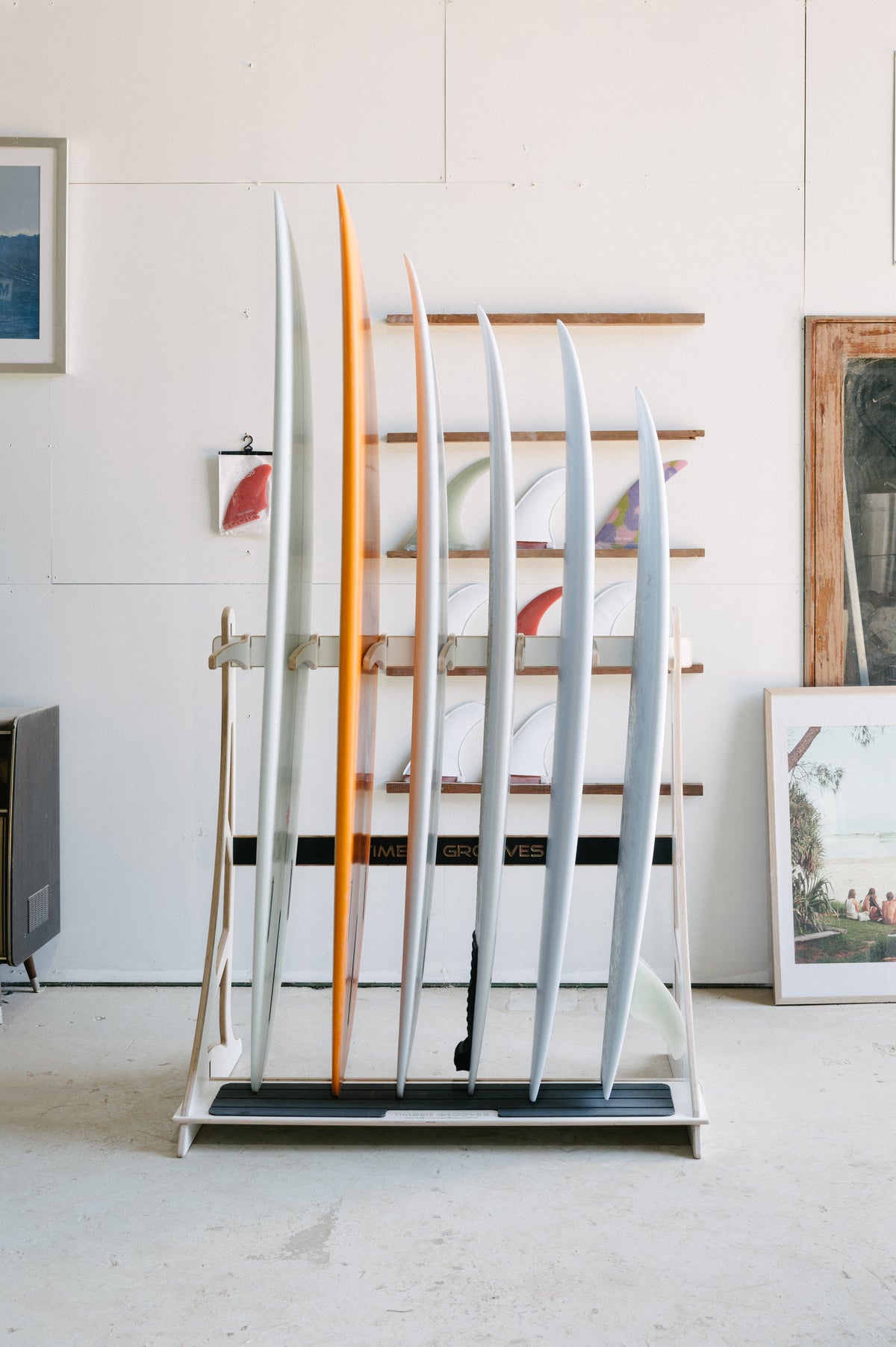 A Timber Grooves Surfboard Rack is mounted on the wall, holding seven surfboards of various sizes and colors, set against a white wall adorned with framed art and photographs.